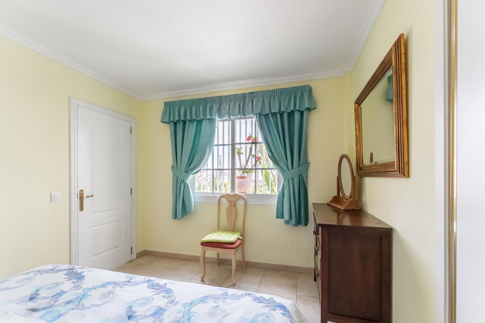A bedroom with a bed, a chair, a dresser, a window with curtains, a door, and a mirror on the wall.