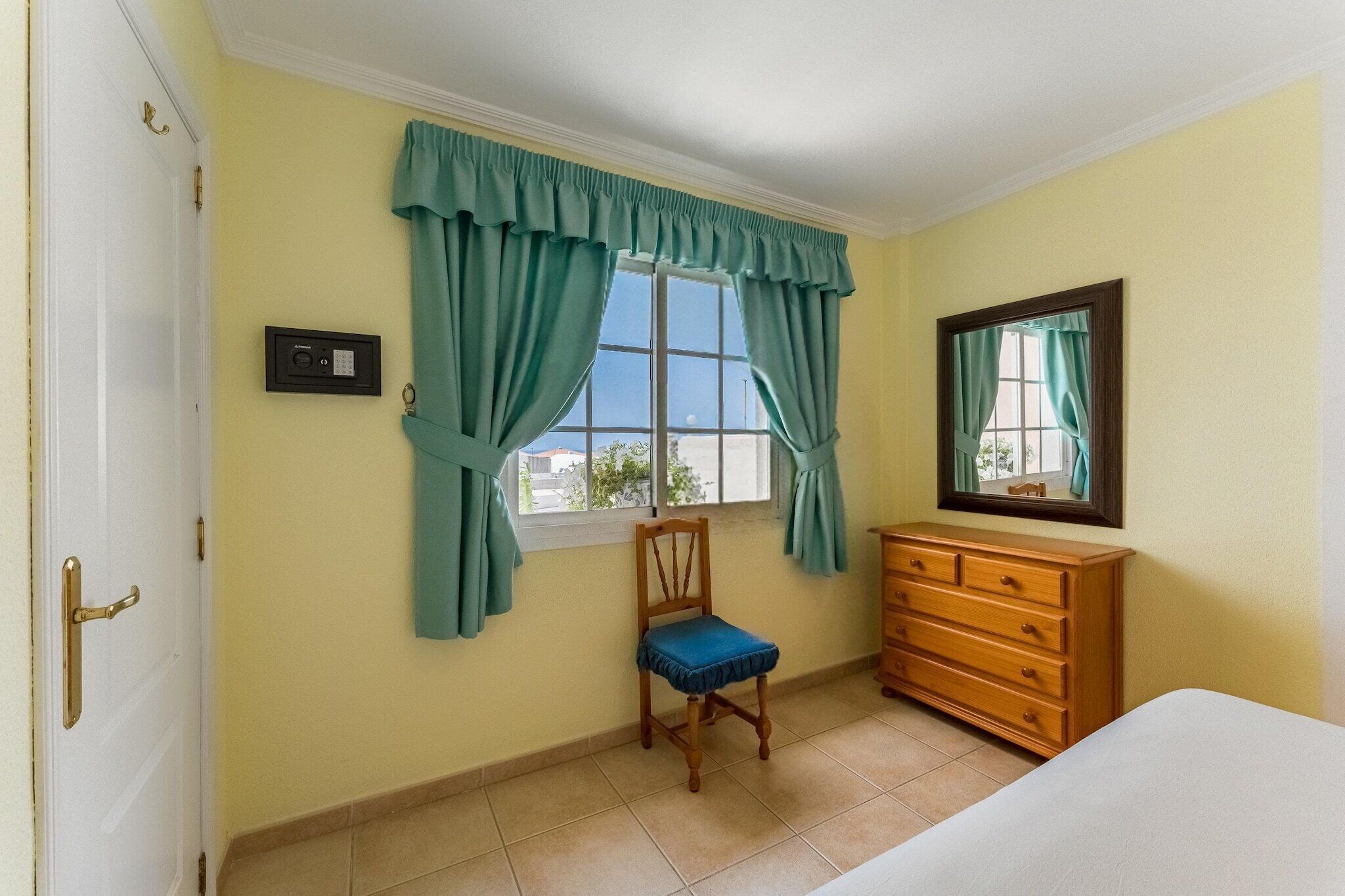Une chambre à coucher comprenant un lit, une commode en bois, une chaise, une fenêtre avec des rideaux et un thermostat fixé au mur.