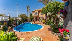A swimming pool with a tiled deck, surrounded by potted plants and a house in the background.