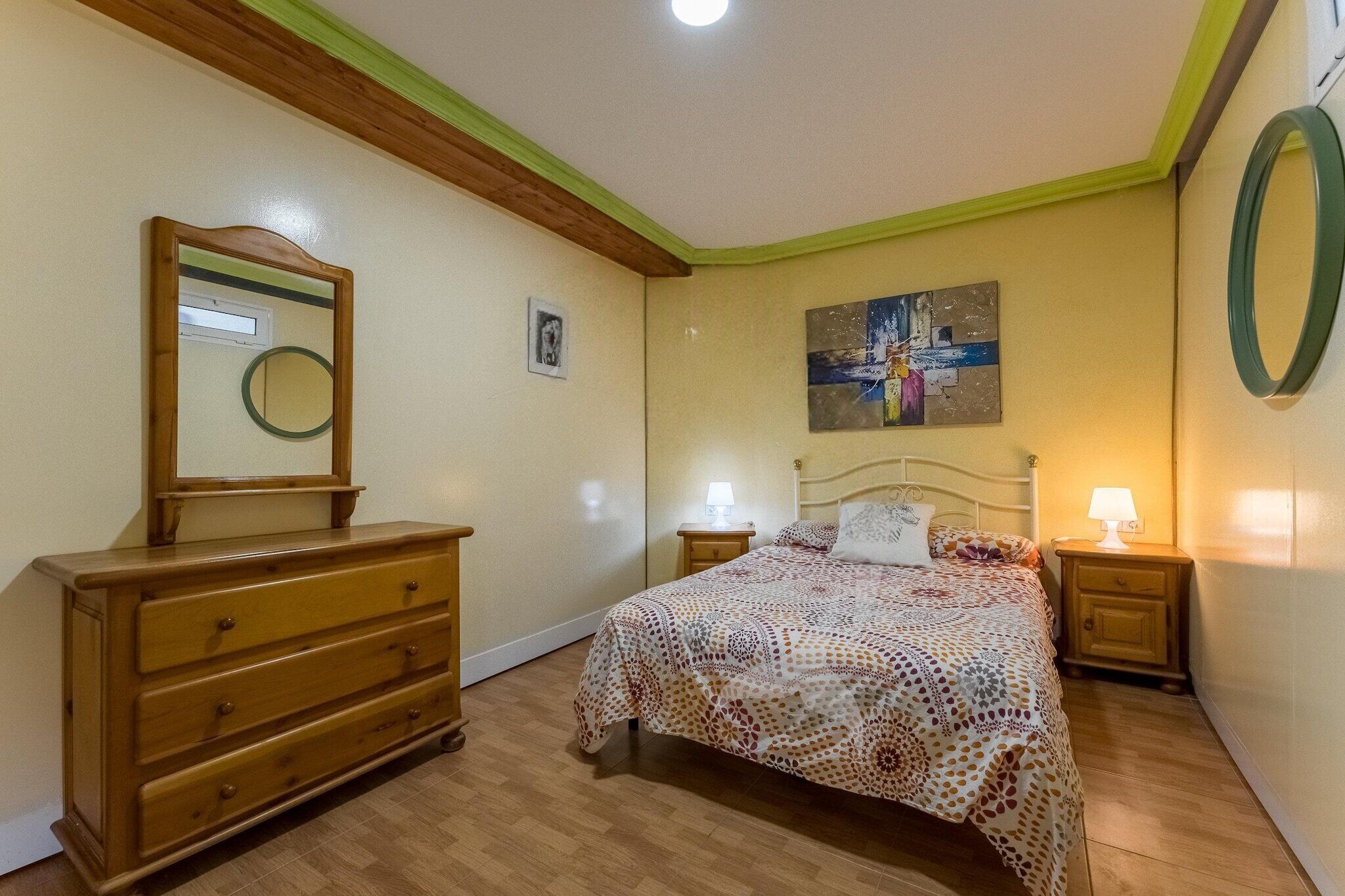A bedroom with a wooden dresser, a bed with a patterned bedspread, two bedside tables with lamps, a wall-mounted mirror, and a painting on the wall.