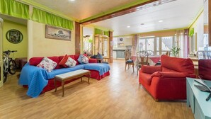Ein geräumiges Wohnzimmer mit einem roten Sofa, einem blauen Sofa und einem Holztisch. Im Hintergrund befindet sich ein Essbereich mit Tisch und Stühlen sowie eine Küche.