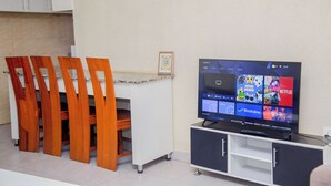 Una sala de estar moderna con una televisión de pantalla plana sobre un mueble, sillas de madera, un pequeño armario y piso de baldosas.