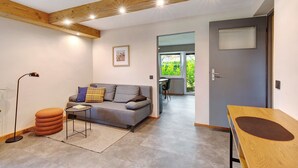 Un salon comprenant un canapé, un pouf et une table, des poutres en bois au plafond et une porte donnant sur un balcon.