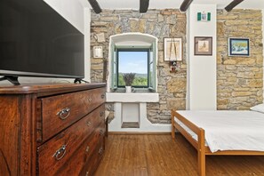 Une chambre avec un mur en pierre, une commode en bois, un lit, une télévision et une fenêtre donnant sur un beau paysage.
