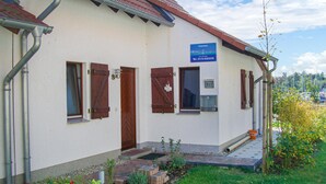 A building with a sign reading "Ferienhaus" and a telephone number. There are three wooden doors and windows, a small garden area with plants, and a paved entrance.