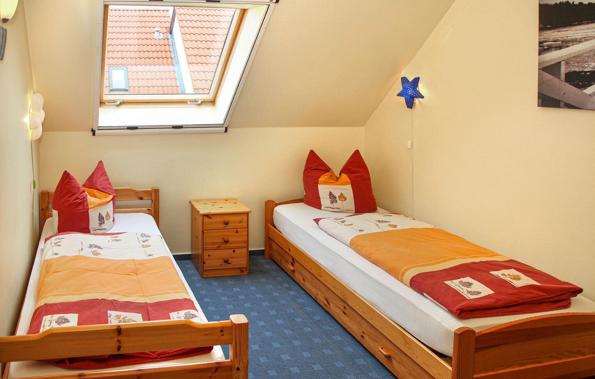 Une petite chambre à coucher accueillante, avec deux lits en bois, une fenêtre donnant sur un toit recouvert de tuiles rouges, une petite table de chevet en bois et une décoration en forme d’étoile bleue sur le mur.