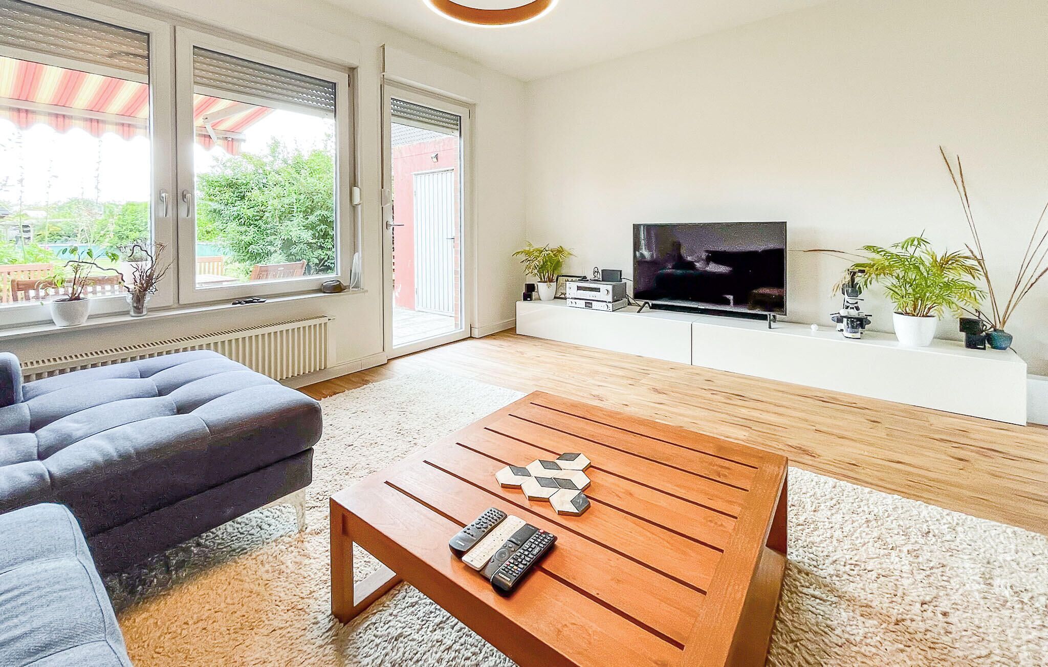 Un soggiorno moderno con un divano grigio, un tavolino in legno con telecomandi, una TV su un mobile bianco e vista su piante verdi all'esterno.