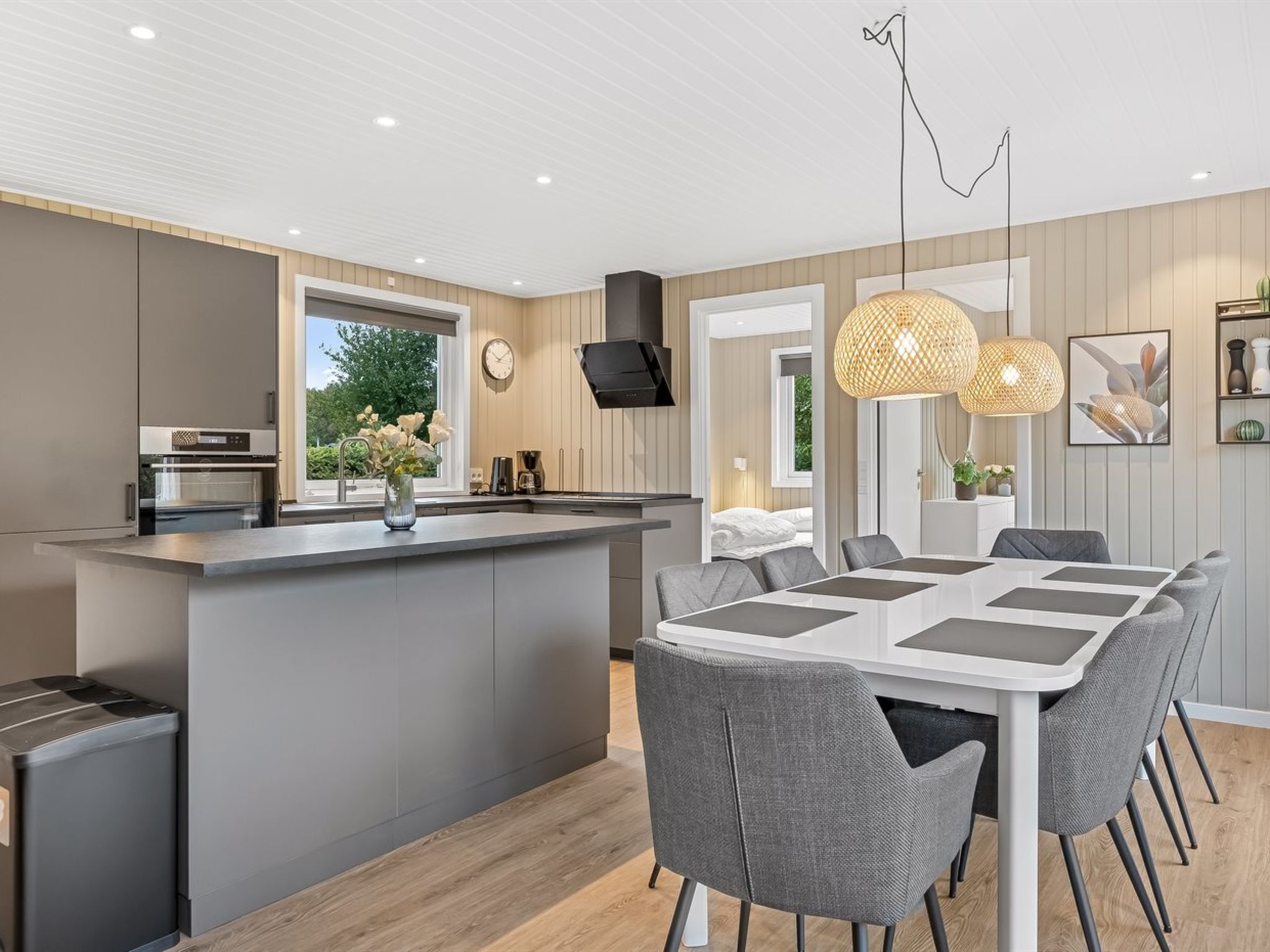 Une cuisine moderne dotée d’un îlot central, un coin repas avec une table blanche et des chaises grises, et un espace de vie comprenant un lit et un téléviseur.