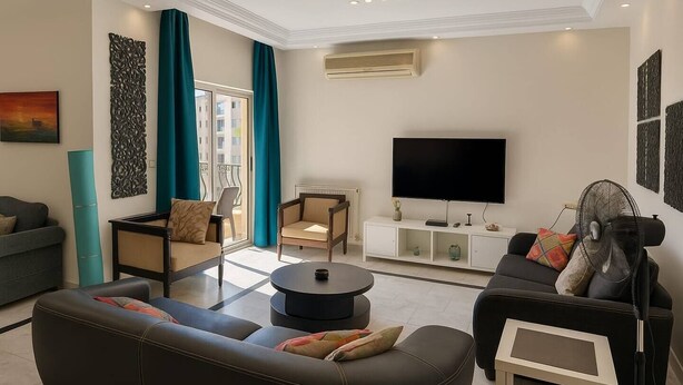 A modern living room with a sofa, armchair, and a TV. There is a small table and a fan. - Central Family Apartment  (Amman)