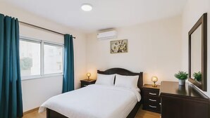 A bedroom with a bed, bedside tables, a dresser, a window with curtains, an air conditioner, and a wall-mounted picture. - Central Family Apartment  (Amman)