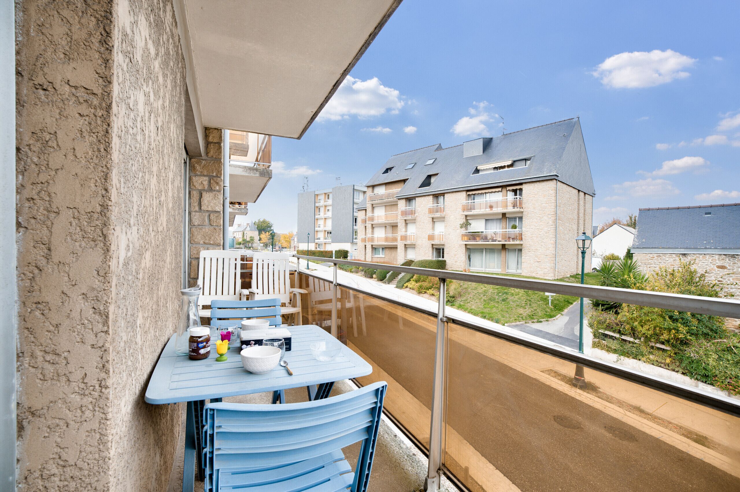 Un balcon avec une table et des chaises, donnant sur une rue bordée d’immeubles et d’espaces verts.