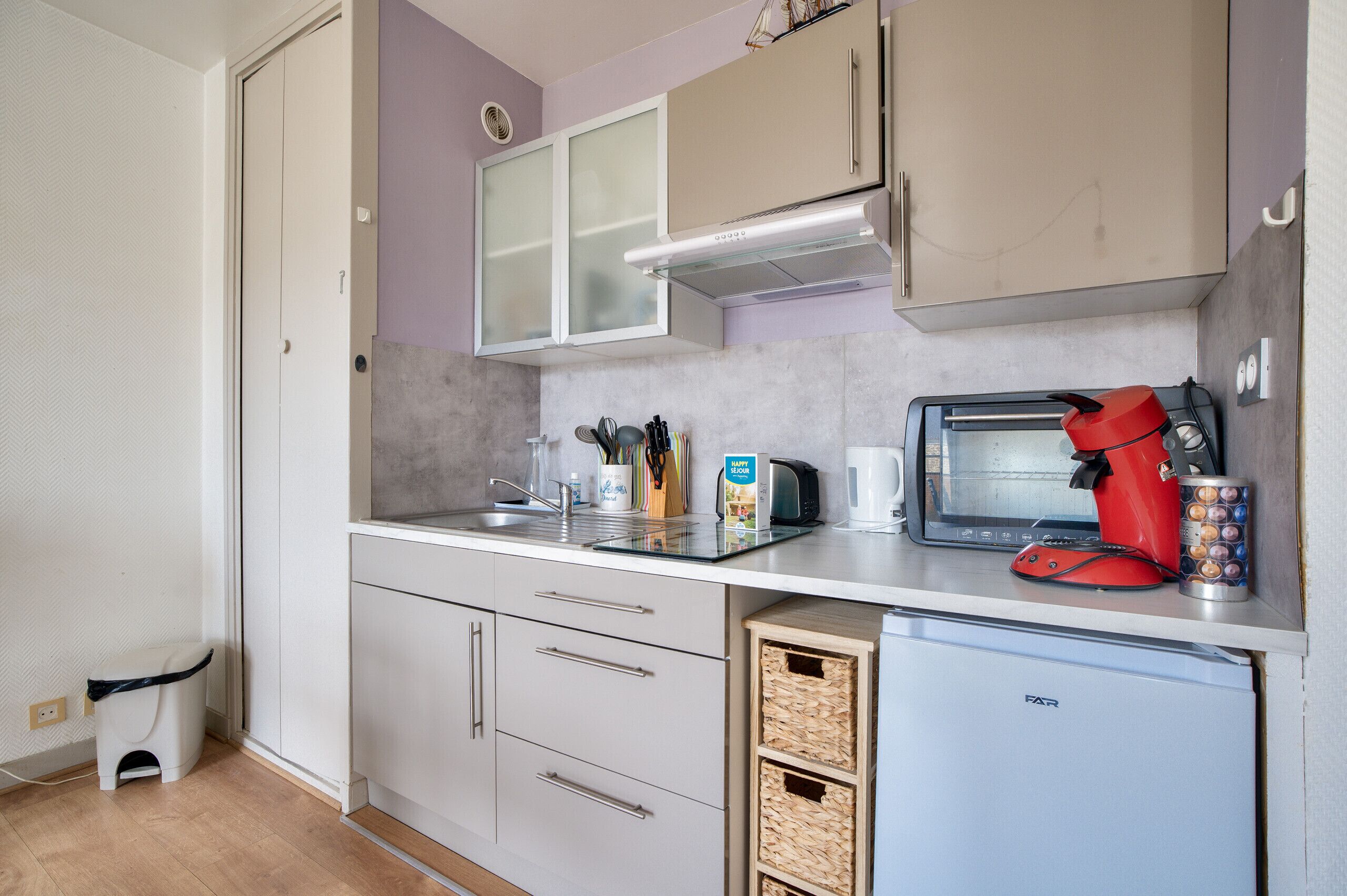 A compact kitchen with a microwave, toaster, and coffee maker on the counter, a small refrigerator, and a trash bin.