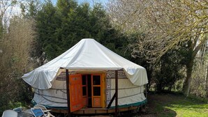 A yurt with an open door, surrounded by a gravel area with a fire pit and wooden stools.