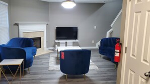Un salón con dos sillones azules, una chimenea blanca, un televisor de pantalla plana y un extintor rojo.