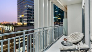 Un balcon donnant sur un paysage urbain, avec notamment un grand immeuble dont les fenêtres sont illuminées. On y trouve deux chaises et une petite table sur laquelle est posé un vase de fleurs.