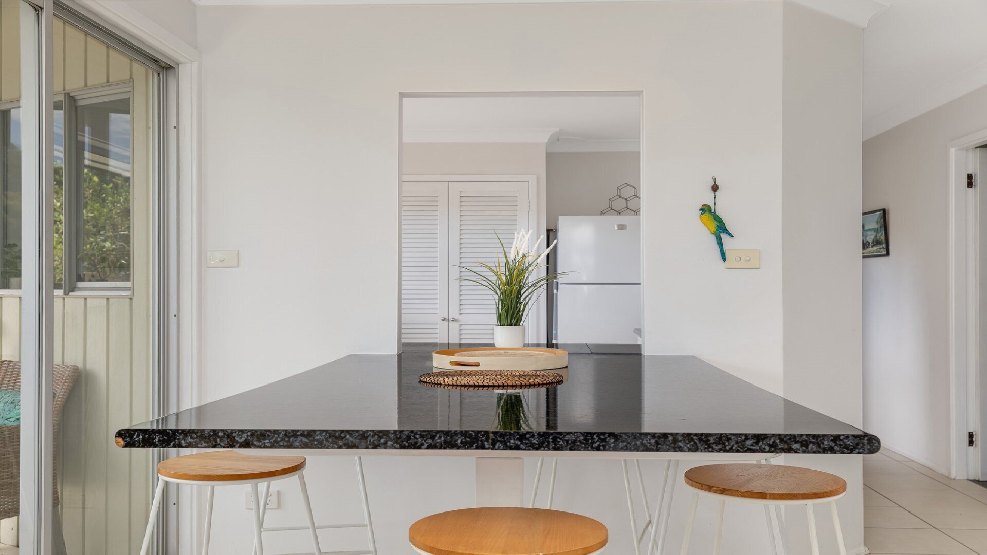 Une cuisine dotée d’un plan de travail en marbre noir, de trois tabourets de bar blancs, d’une planche à découper en bois, d’une plante en pot et d’un ornement en forme d’oiseau suspendu.