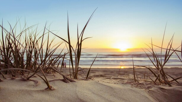 A beach scene with sand and grass in the foreground and a sunset over the ocean in the background.