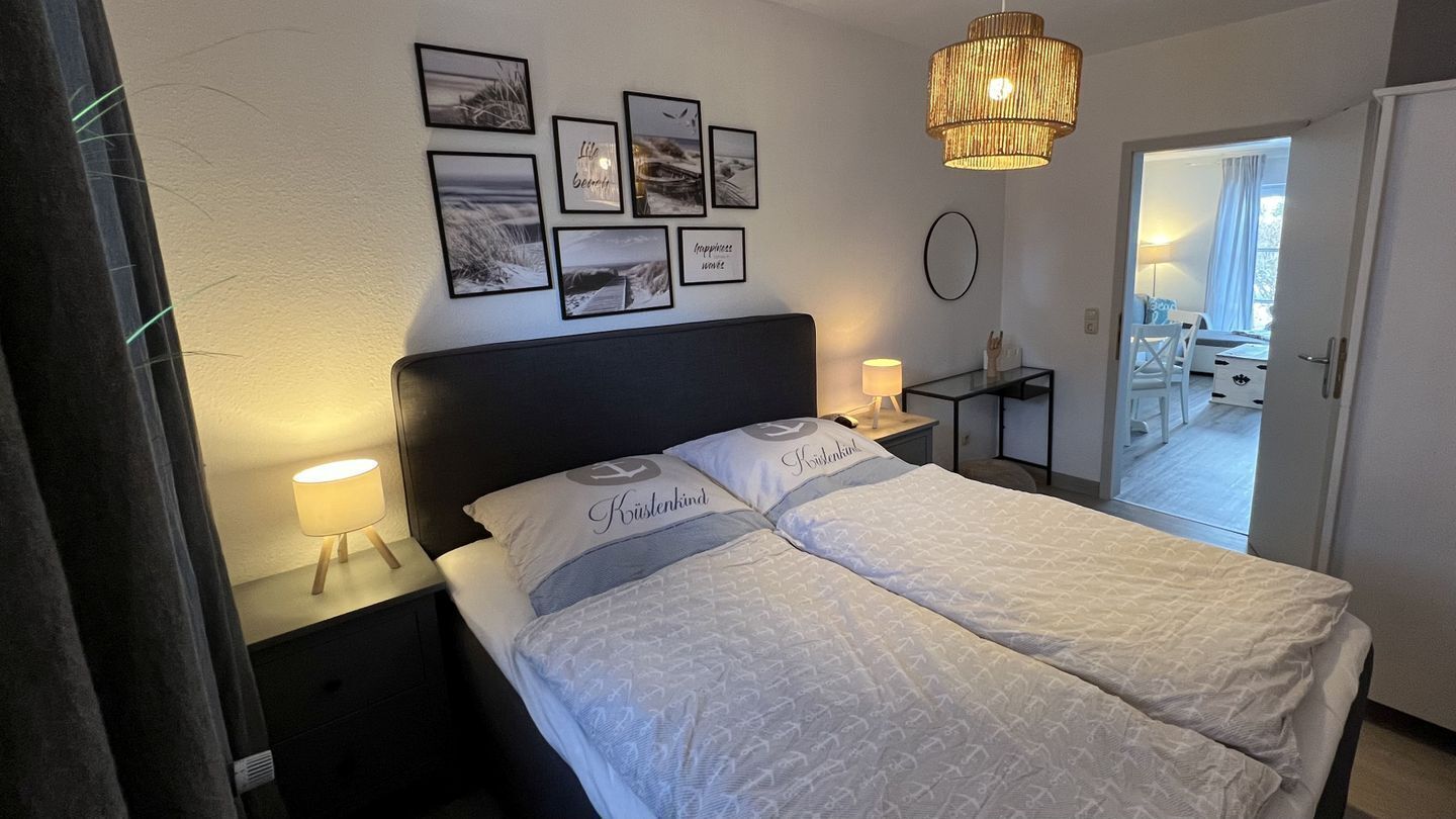 A neatly arranged bedroom with a bed, bedside lamps, a bedside table, and a wall with framed pictures.