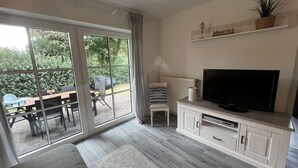Una sala de estar con televisor, muebles blancos, un sofá y un ventanal corredizo que da a un área exterior con una mesa y sillas.