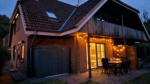 Una casa de dos pisos con techo inclinado, un porche cubierto con asientos exteriores y guirnaldas de luces.