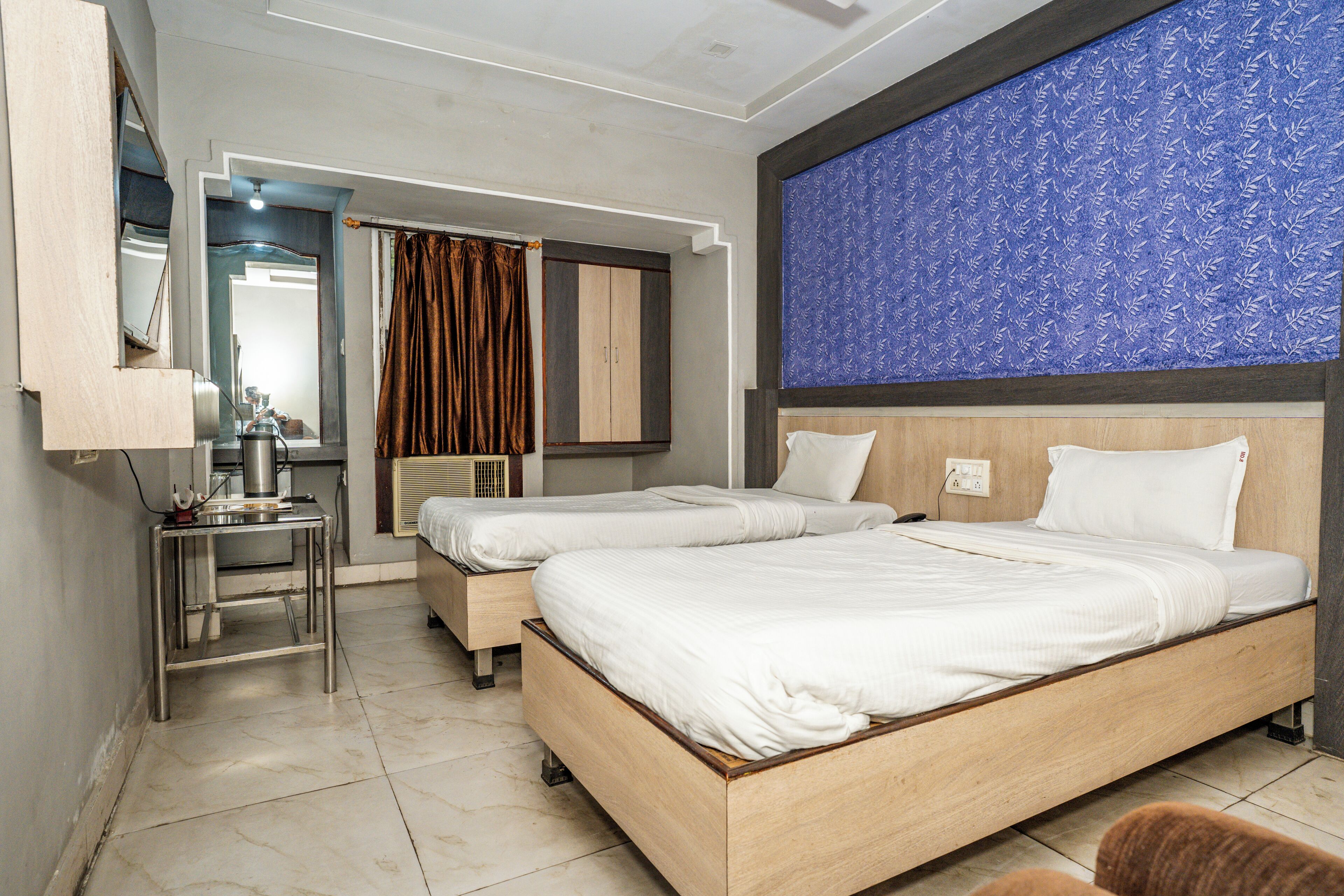 A hotel room with two beds, a small table, a mirror, and a blue patterned wall.