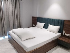 A neatly made bed with white linens in a modern room with a wooden headboard and a grey curtain. - Hotel Sun-Om (Raipur)