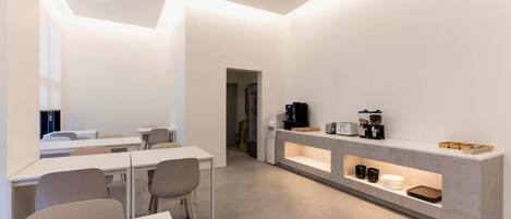 Una cafetería moderna y minimalista con paredes blancas, una barra larga con máquinas de café y zonas de estar con mesas y sillas.
