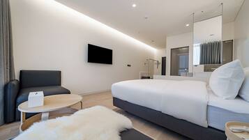 A modern hotel room with a large bed, a flat-screen TV mounted on the wall, a seating area with a chair and a small table, and a large mirror above the bed.