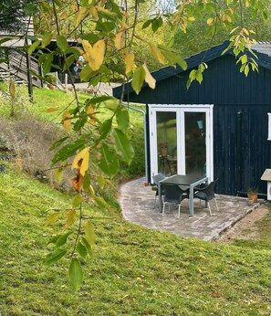 Un espace extérieur abrité, avec une table et des chaises, une plante en pot, et un bâtiment de couleur sombre en arrière-plan.