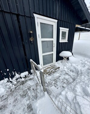 L’extérieur d’un bâtiment est recouvert de neige. On y voit une porte à cadre blanc, une petite fenêtre et un banc également couvert de neige.