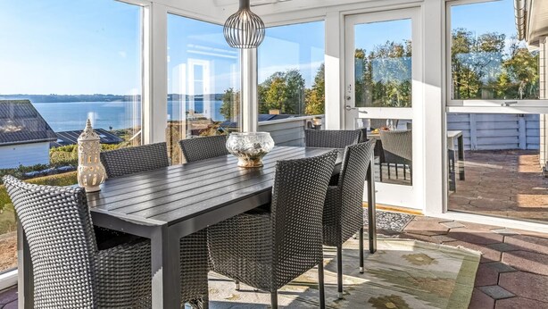 Un patio iluminado por el sol con una mesa y sillas, amplias ventanas que ofrecen vista al mar, y una pérgola en lo alto.