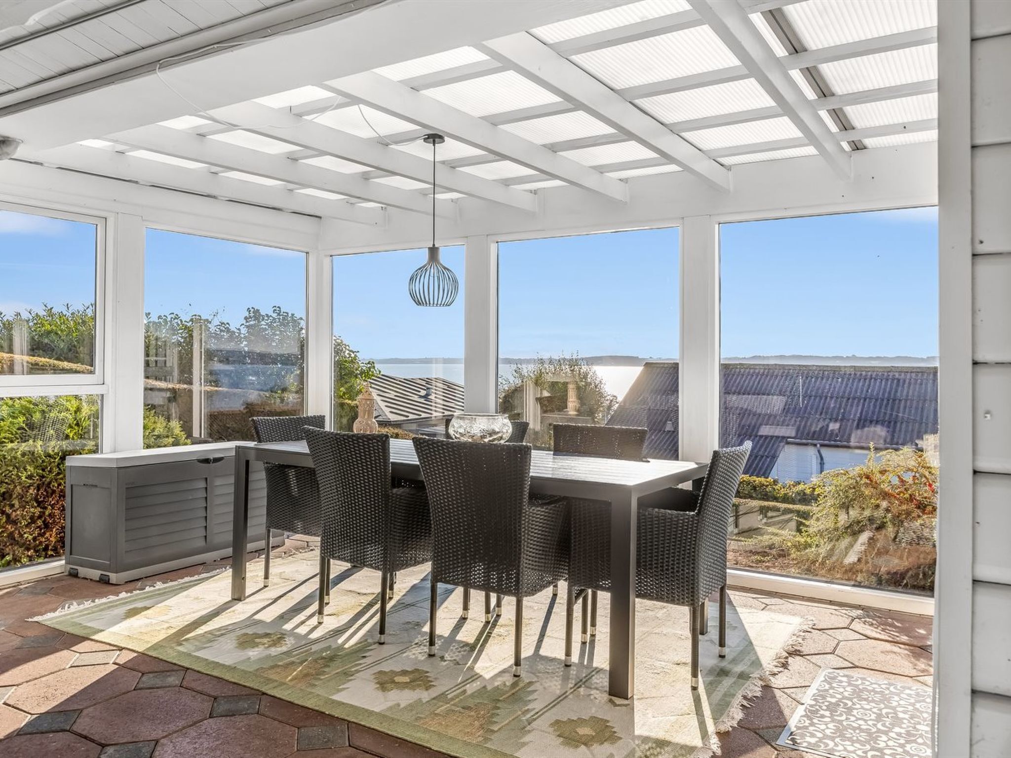 Un patio cubierto con mesa y sillas, ventanas amplias y vista al mar.