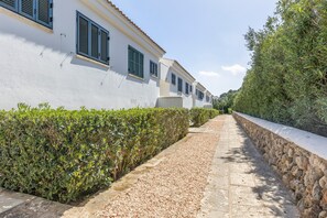 A pathway flanked by a white building with shutters, a stone wall, and dense greenery.