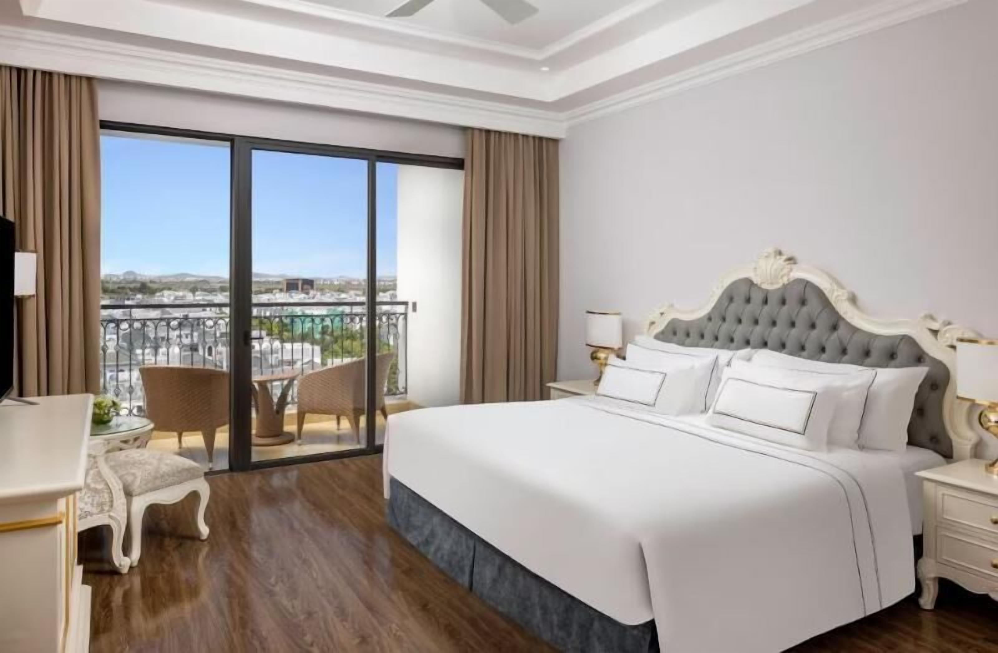 A hotel room with a large bed, a small table, a chair, a nightstand, and a view of a balcony with a cityscape.
