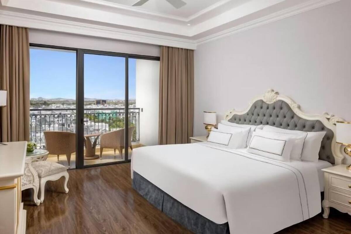 Quarto de hotel com uma cama grande, uma mesa pequena, uma cadeira, uma mesinha de cabeceira e vista para uma varanda com panorama da cidade.