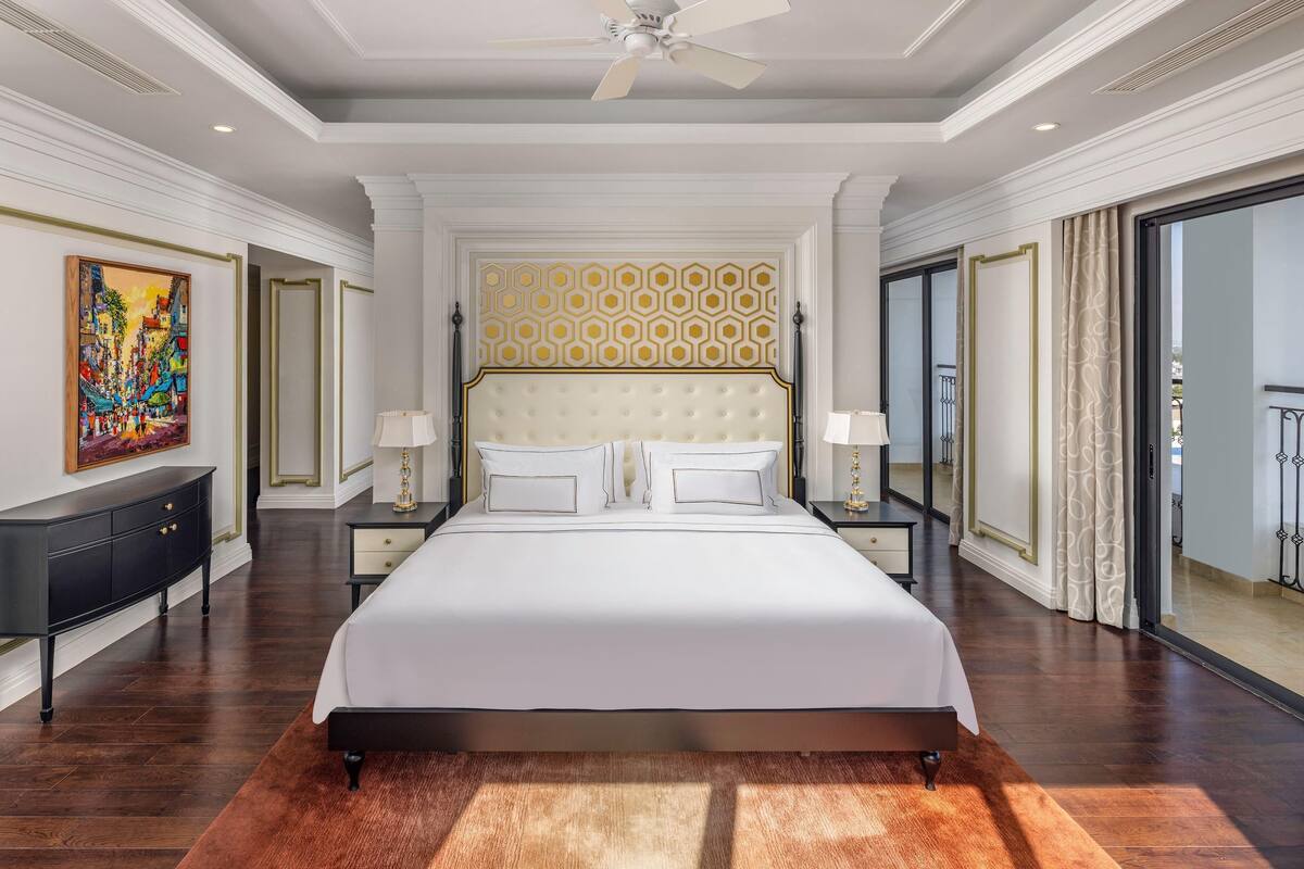 Uma cama grande com cabeceira estofada, luminárias de cabeceira, um quadro na parede e vista através de uma porta de vidro.