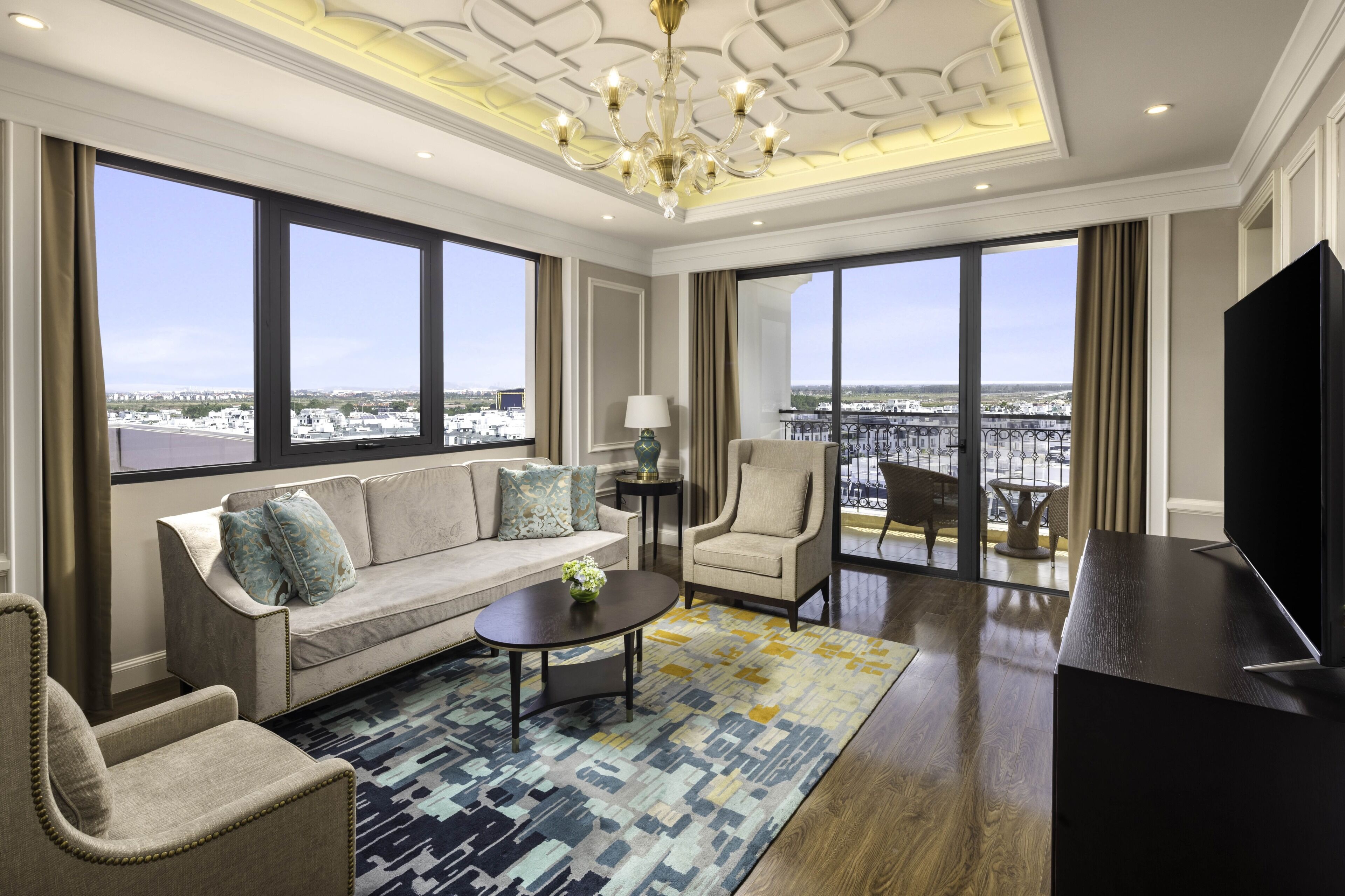 A spacious living room with a sofa, armchairs, a coffee table, a TV, and large windows offering a city view.