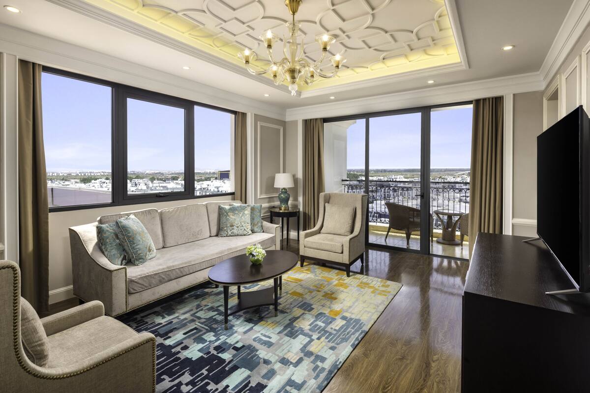 Uma sala de estar ampla com sofá, poltronas, mesa de centro, televisão e janelas grandes com vista para a cidade.