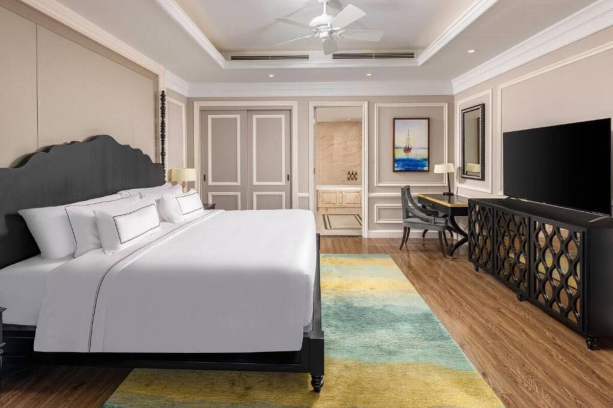 Quarto de hotel com uma cama grande, uma escrivaninha com cadeira, televisão e ventilador de teto.