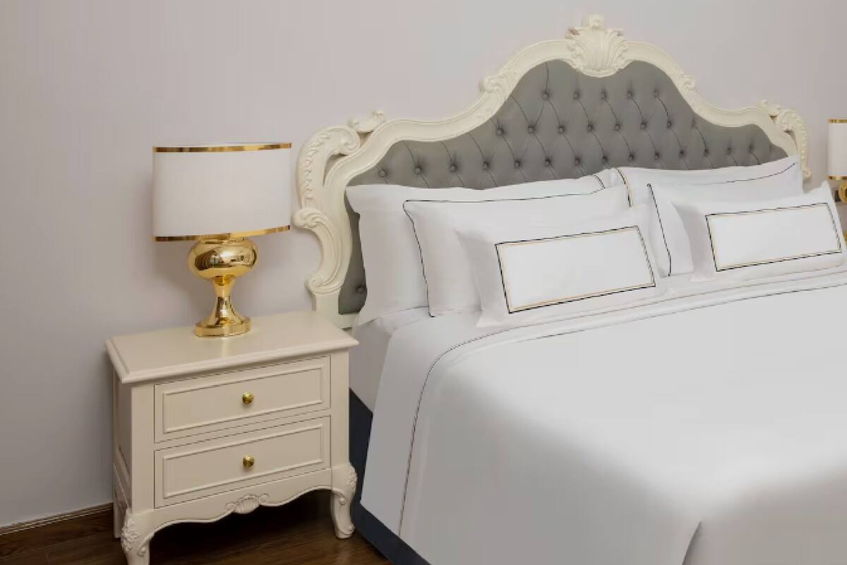 Uma cama com cabeceira estofada, roupa de cama branca e abajur dourado em uma mesa de cabeceira.