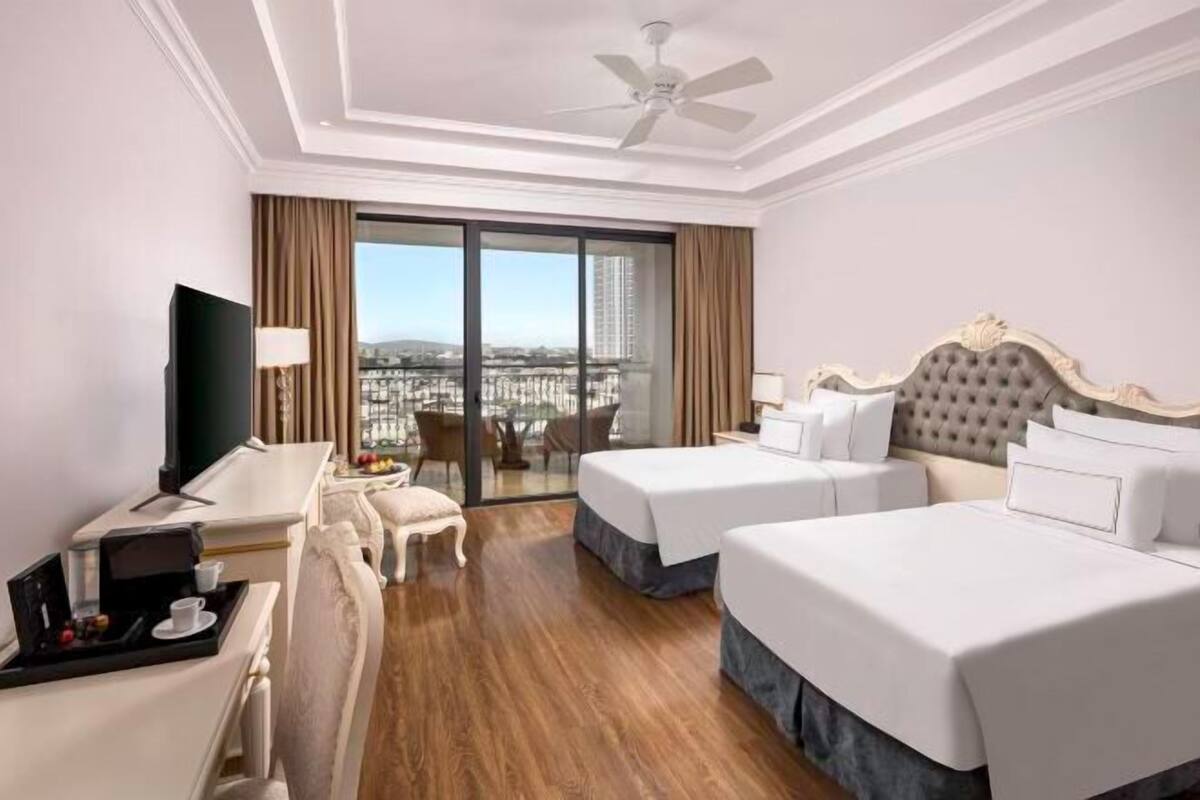 Quarto de hotel com duas camas, televisão, escrivaninha com bandeja, cadeira e varanda com vista para a cidade.