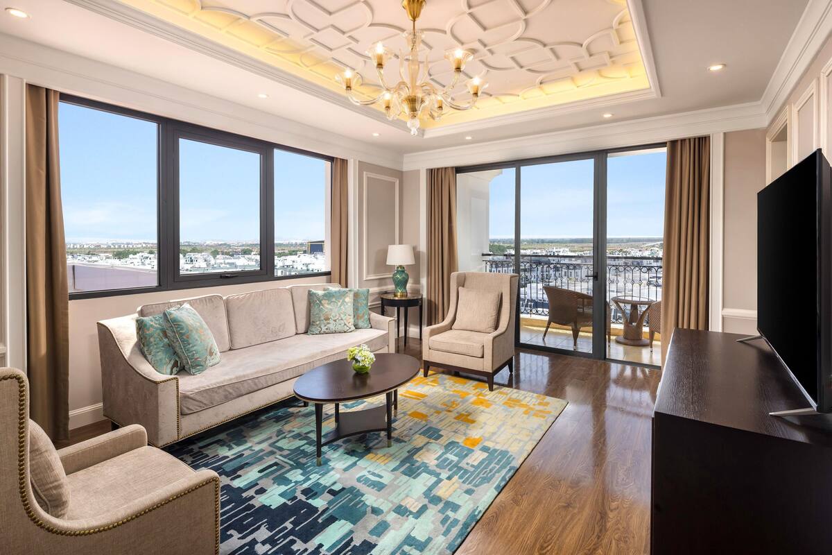 Uma sala de estar espaçosa com sofá, poltronas, mesa de centro e televisão. Grandes janelas proporcionam vista para a cidade.