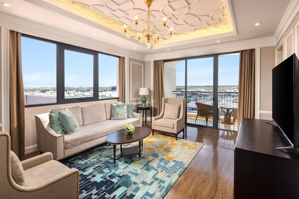 A spacious living room with a sofa, armchairs, a coffee table, and a TV. Large windows offer a city view.