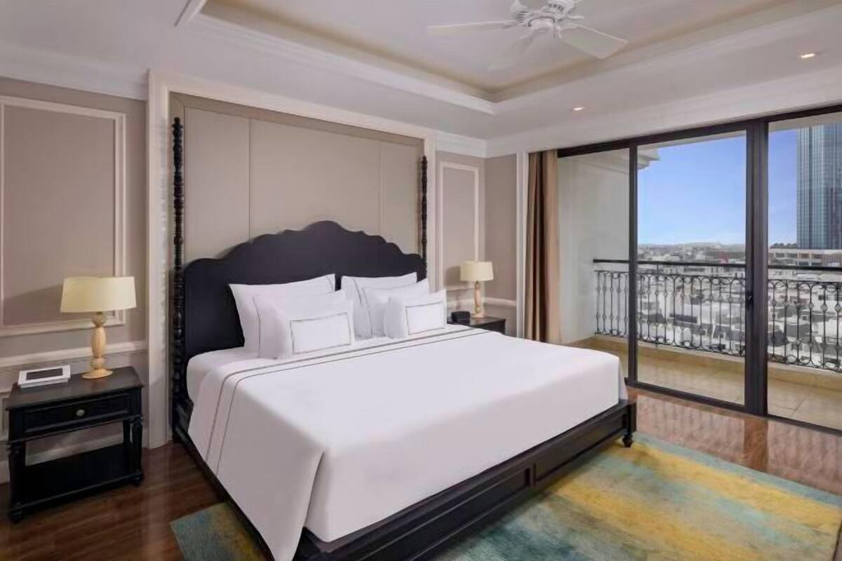 Quarto de hotel com uma cama grande, duas mesas de cabeceira com luminárias, um ventilador de teto e uma varanda com vista para a cidade.