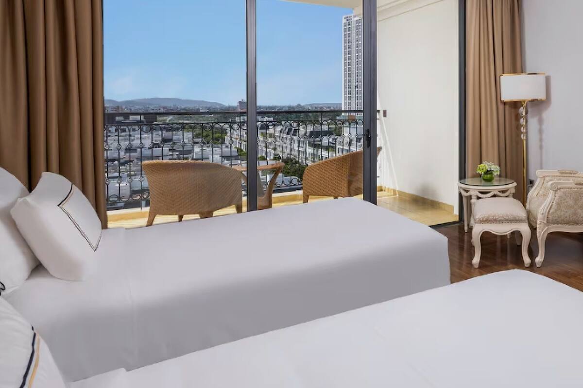 Quarto de hotel com uma cama grande, duas cadeiras, uma mesa pequena e vista para a paisagem urbana através de grandes janelas.