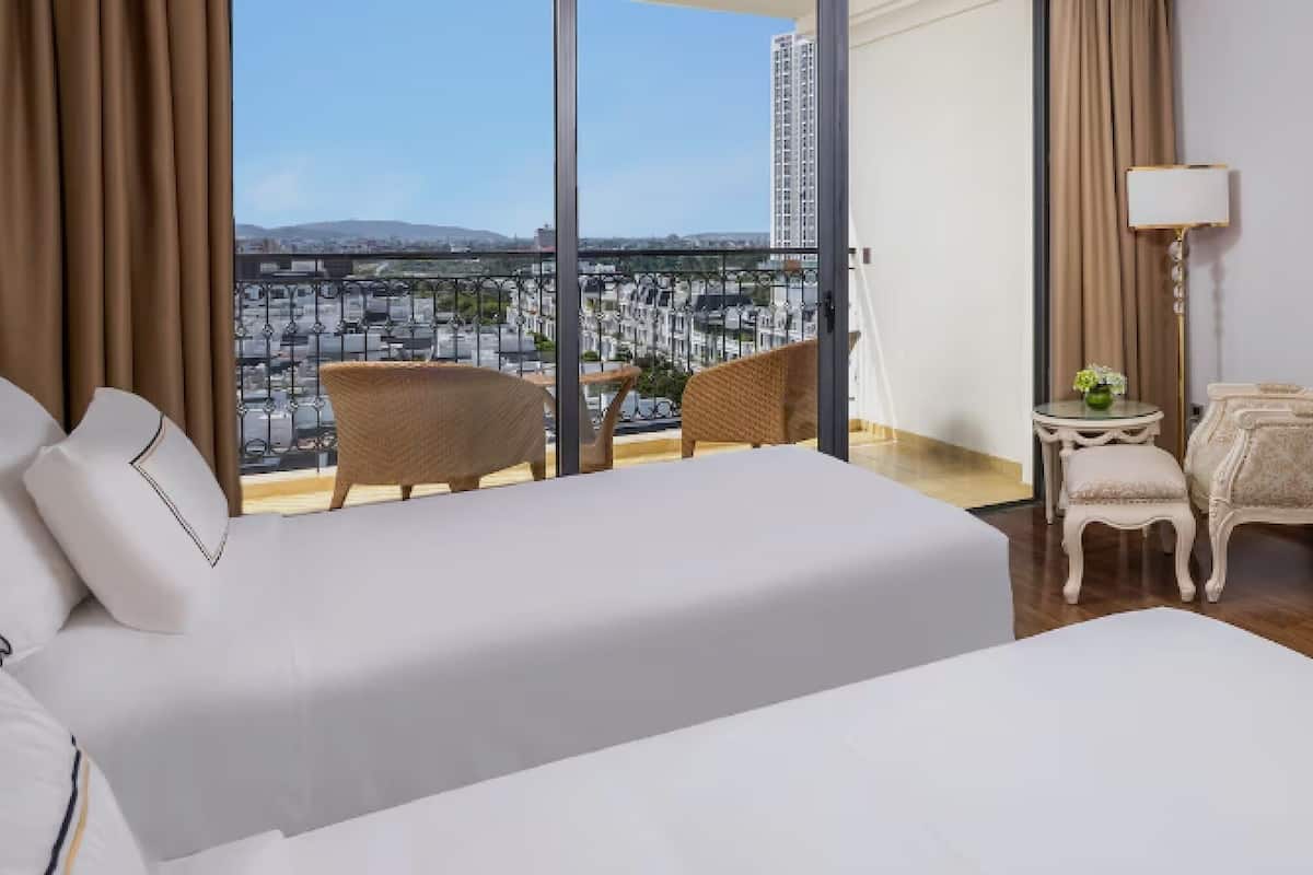A hotel room with a large bed, two chairs, a small table, and a view of a cityscape through large windows.