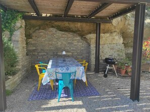 A covered outdoor seating area with a stone wall backdrop, a table with a patterned tablecloth, and colorful chairs.
