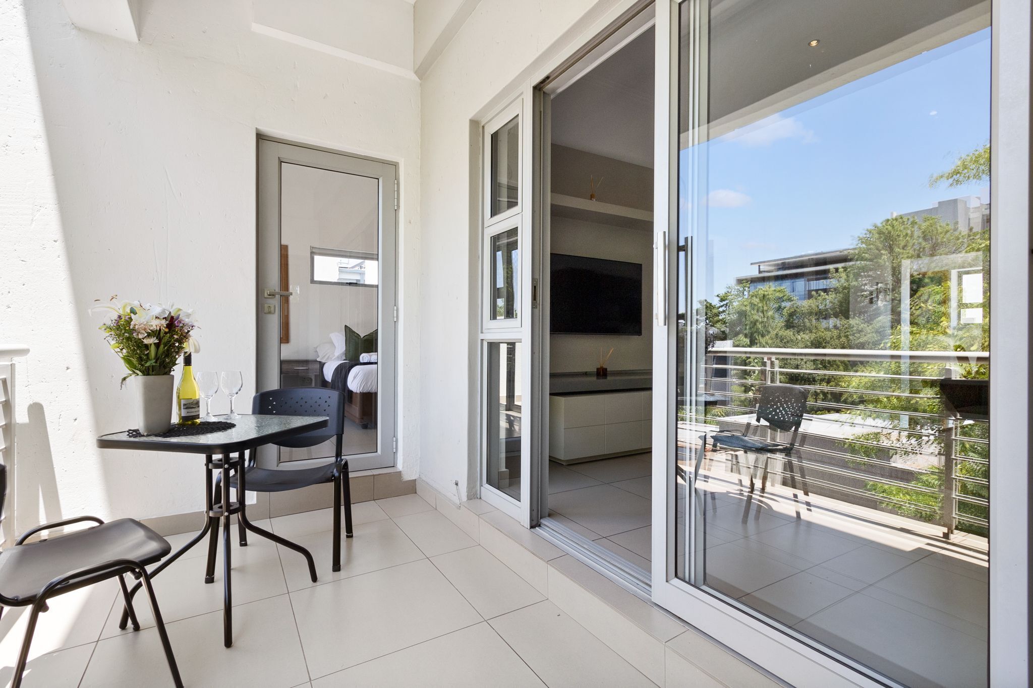 A balcony with a dining area, a sliding glass door leading to a kitchen, and a view of the surrounding buildings.