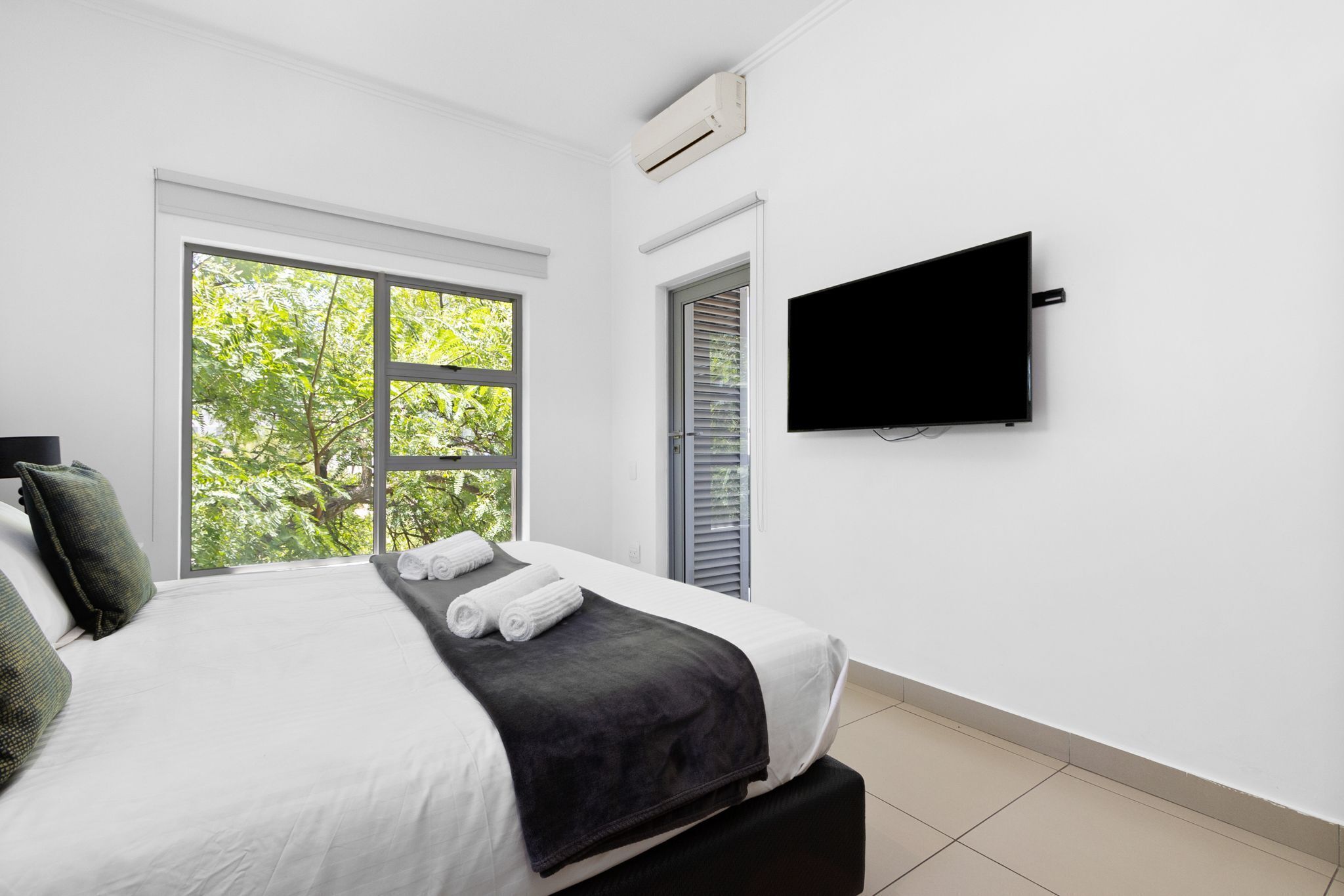 A modern bedroom with a large window, a flat-screen TV, and a neatly made bed.
