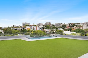 Eine Dachterrasse mit Schwimmbad, umgeben von einem gepflegten Rasen und Blick auf eine Stadt mit verschiedenen Gebäuden.